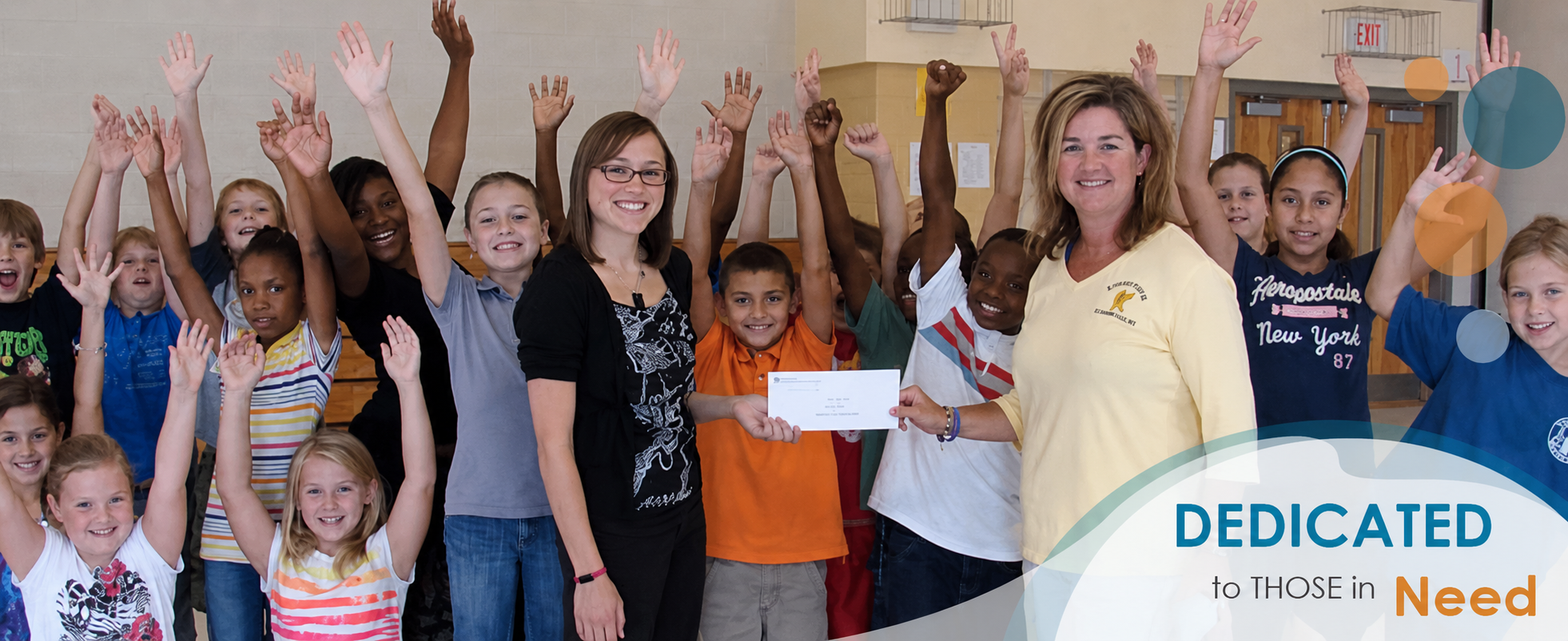 Children and teachers indoors with hands raised, one holding a presentation envelope
